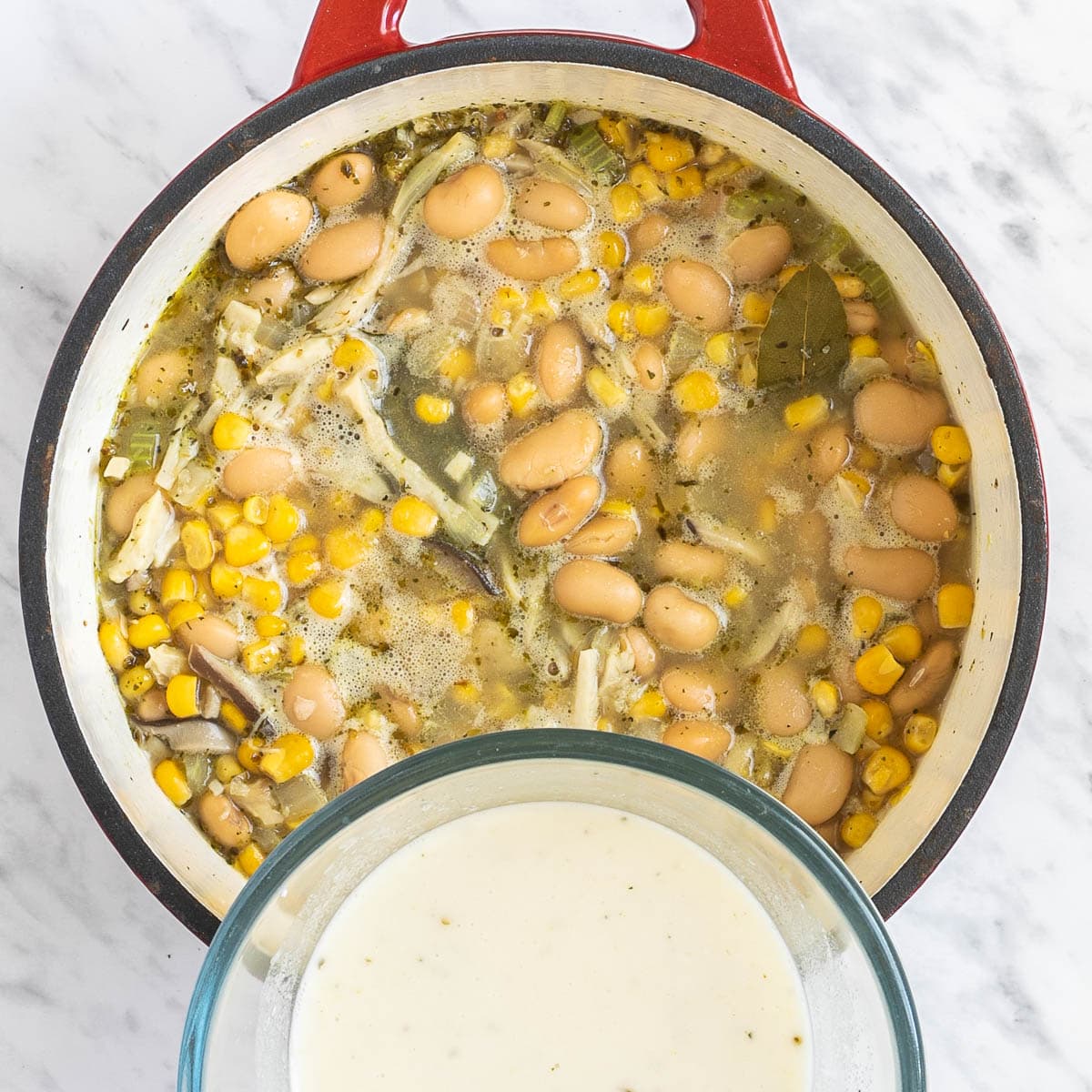 Red Dutch oven with shredded mushrooms, white beans, and sweet corns in a thin light green white soup. A hand is holding a small bowl of white sauce.