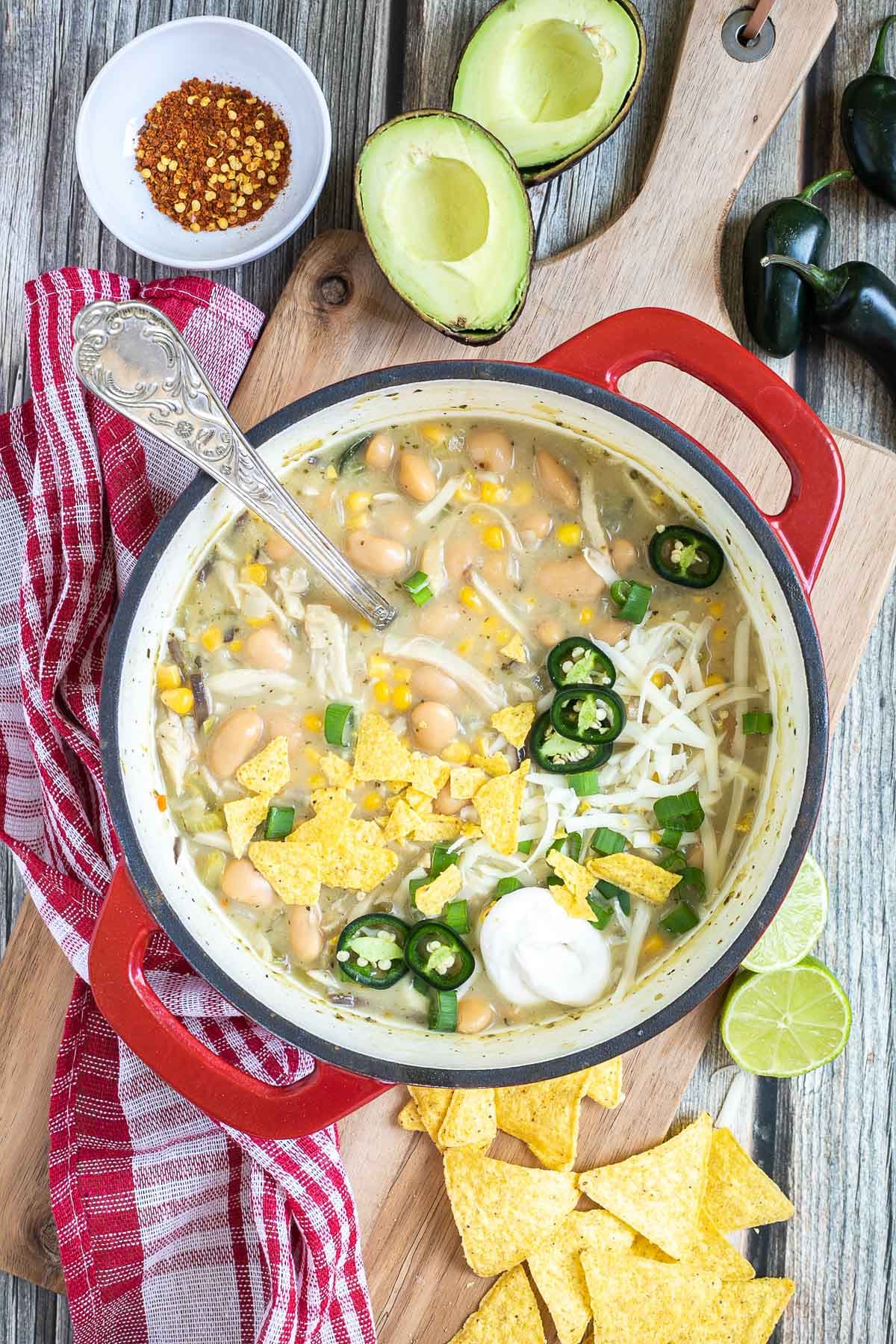 A red enameled Dutch oven with a light brown chili with white beans, shredded mushrooms, corn, topped with green chili slices, shredded cheese and nacho chip pieces. Avocado, and other toppings are around it.
