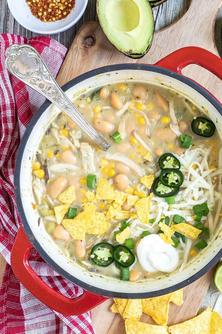 A red enameled Dutch oven with a light brown chili with white beans, shredded mushrooms, corn, topped with green chili slices, shredded cheese and nacho chip pieces. Avocado, and other toppings are around it.