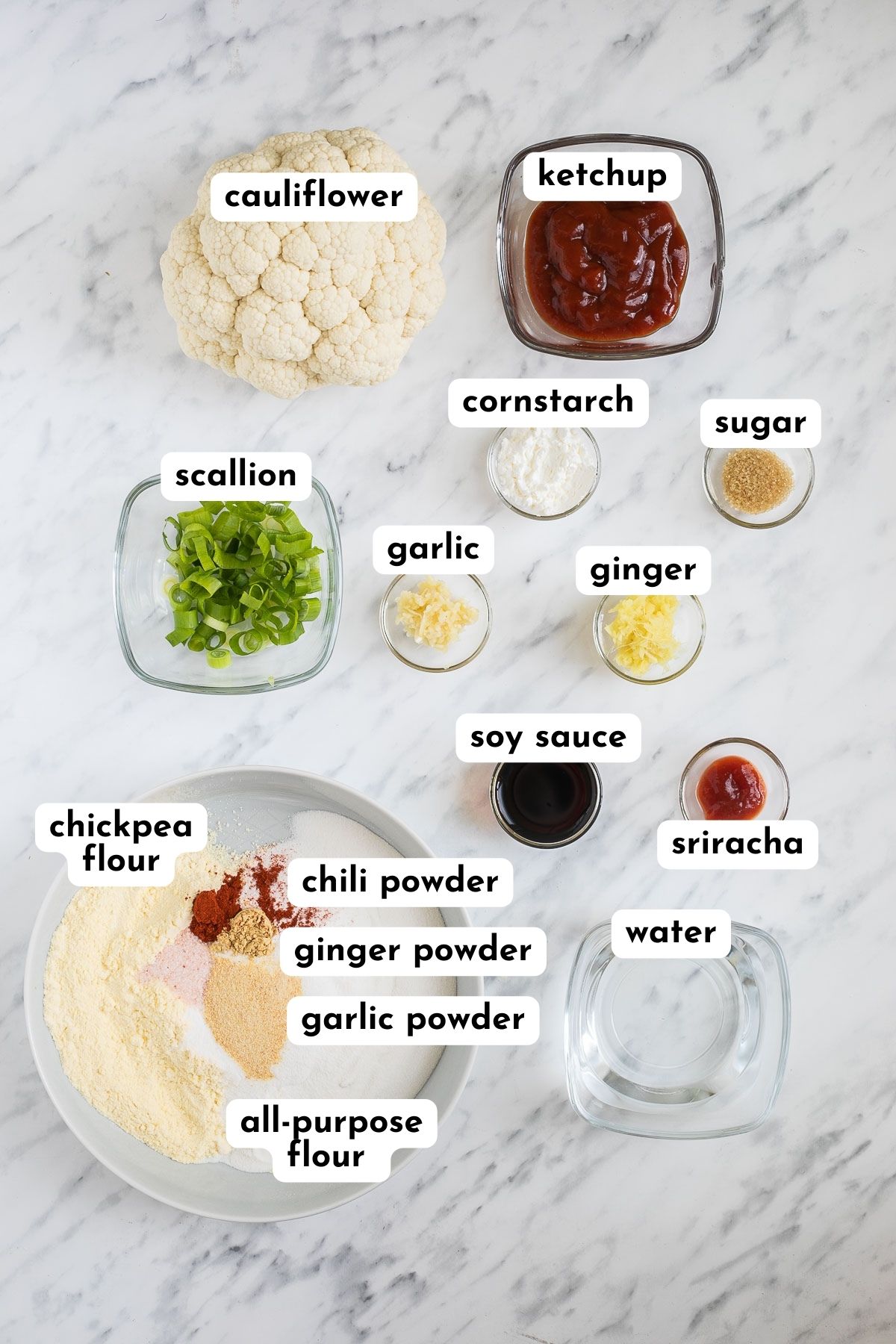 The ingredients of gobi manchurian in glass bowls like scallion, garlic, ginger, soy sauce, sriracha, water, starch, and ketchup. There is a whole head of cauliflower next to it and a white bowl with white and yellow flour and red to yellow spices.