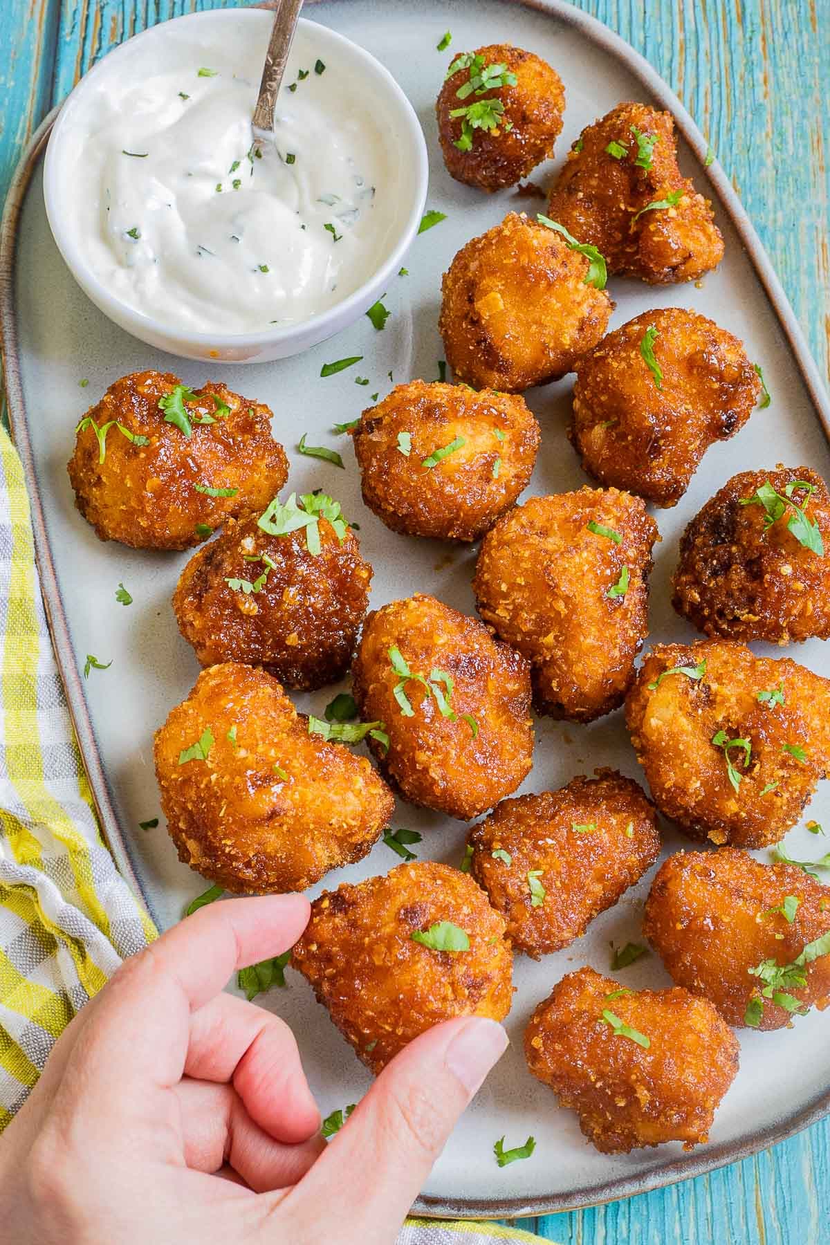 Breaded cauliflower florets in a light blue plate sprinkled with chopped green herbs and served with a small bowl of white sauce. A hand is taking one.
