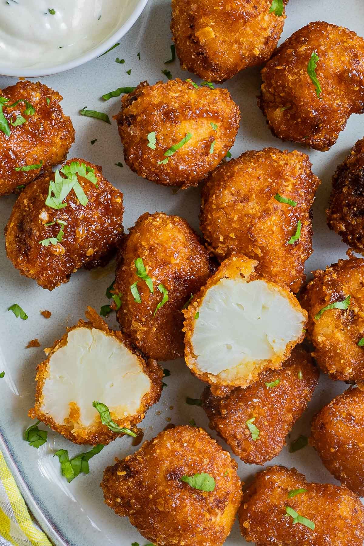 Breaded cauliflower florets in a light blue plate sprinkled with chopped green herbs and served with a small bowl of white sauce. One floret is cut in half to show the white insides.