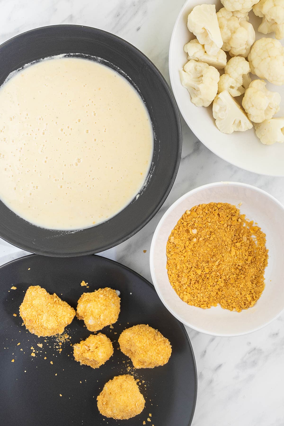 1 bowl with precooked cauliflower florets, one bowl with light wet batter, one bowl with crushed corn flakes and one black plate with some breaded cauliflower