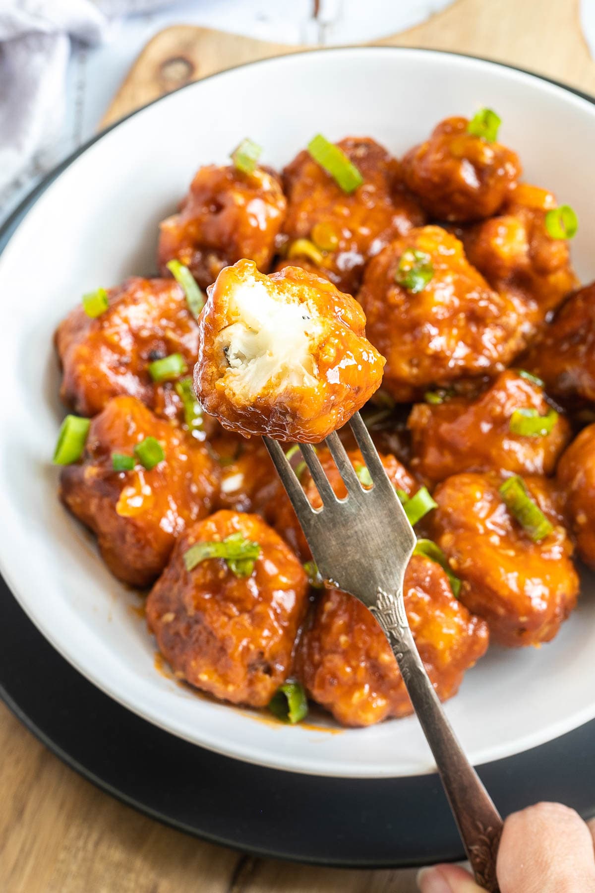 Orange sauce glazed cauliflower florets sprinkled with chopped green scallions in a white plate. A fork is taking one floret and there is a bite size piece missing from it.