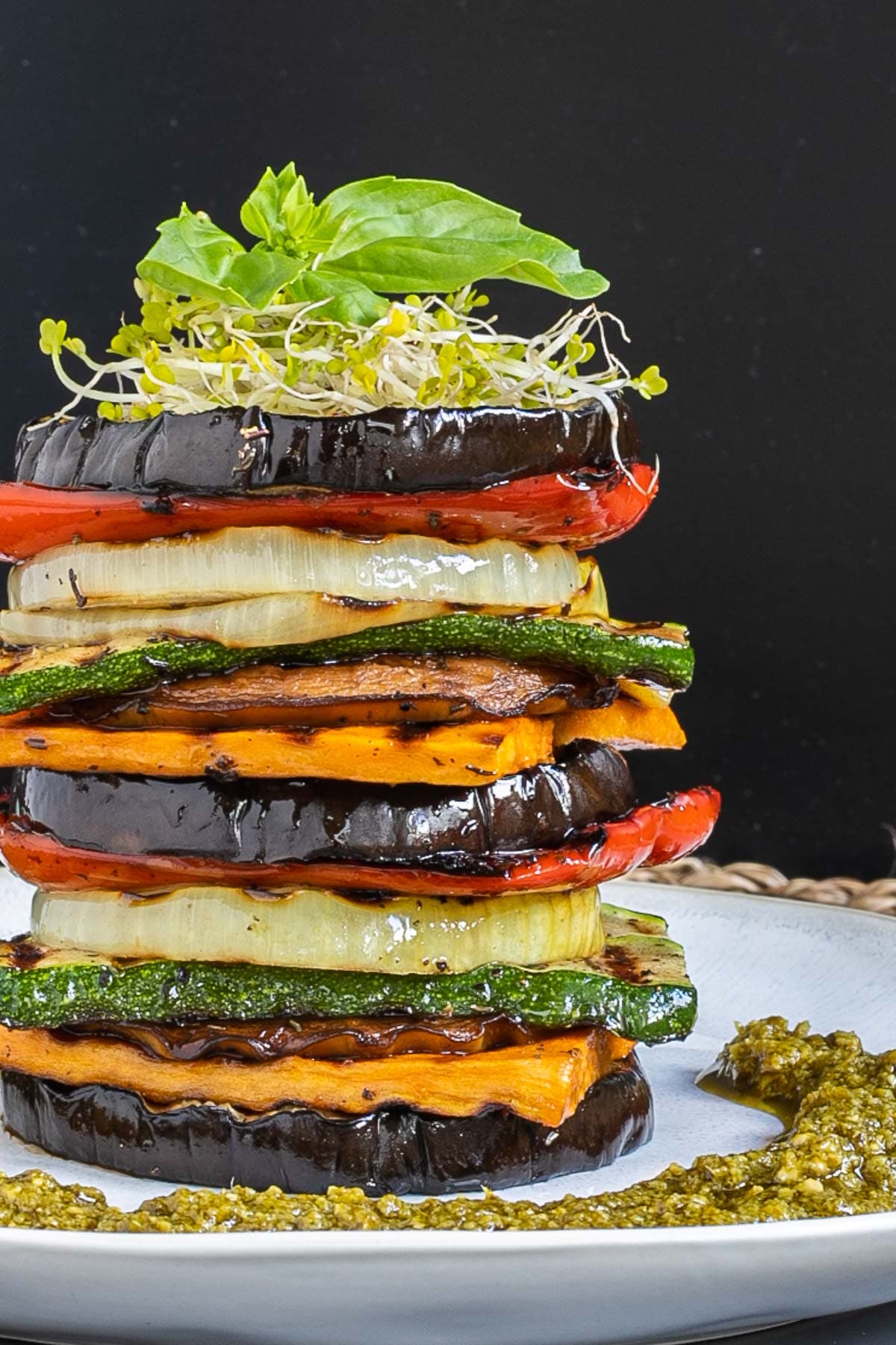 Different veggie slices are stack on top of each other to create a tower of veggies. It has eggplant, zucchini, sweet potato, onion, red bell pepper and mushroom slices. Green pesto is served around it. It is topped with sprouts and basil leaves.