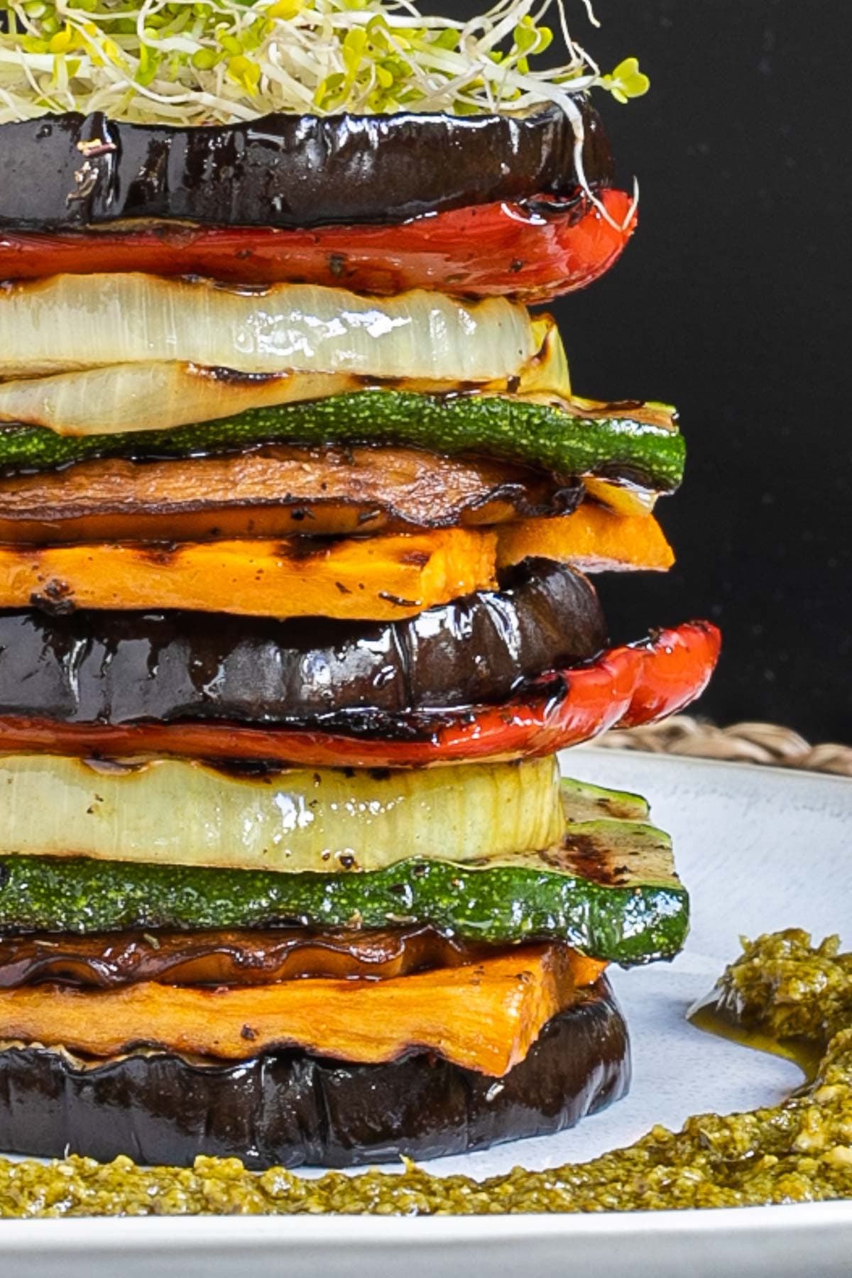 Different veggie slices are stack on top of each other to create a tower of veggies. It has eggplant, zucchini, sweet potato, onion, red bell pepper and mushroom slices. Green pesto is served around it. It is topped with sprouts and basil leaves.