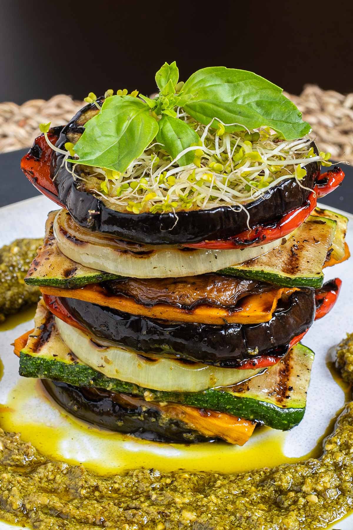 Different veggie slices are stack on top of each other to create a tower of veggies. It has eggplant, zucchini, sweet potato, onion, red bell pepper and mushroom slices. Green pesto is served around it. It is topped with sprouts and basil leaves.