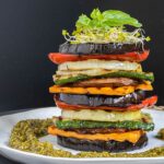 Different veggie slices are stack on top of each other to create a tower of veggies. It has eggplant, zucchini, sweet potato, onion, red bell pepper and mushroom slices. Green pesto is served around it. It is topped with sprouts and basil leaves.