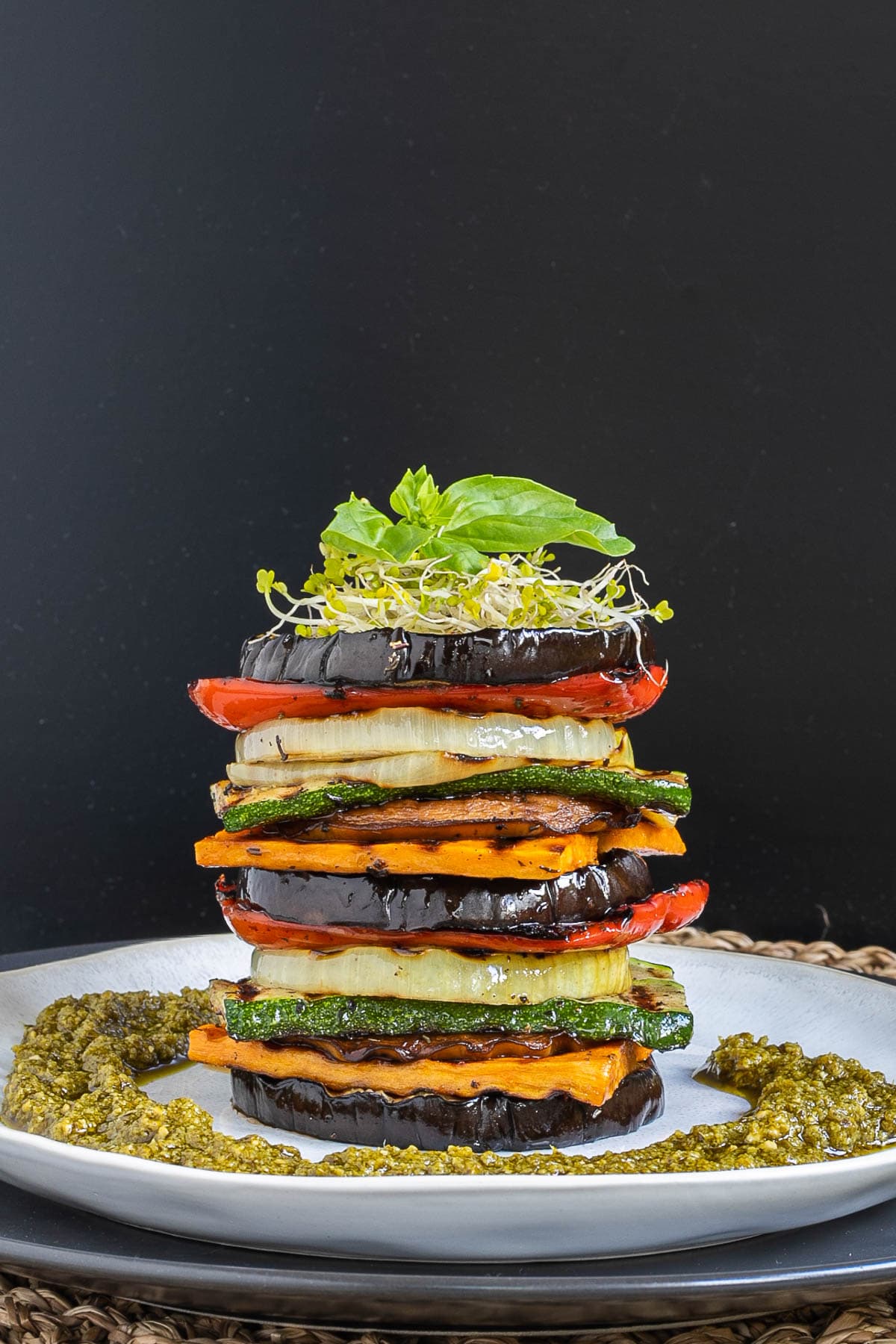 Different veggie slices are stack on top of each other to create a tower of veggies. It has eggplant, zucchini, sweet potato, onion, red bell pepper and mushroom slices. Green pesto is served around it. It is topped with sprouts and basil leaves.
