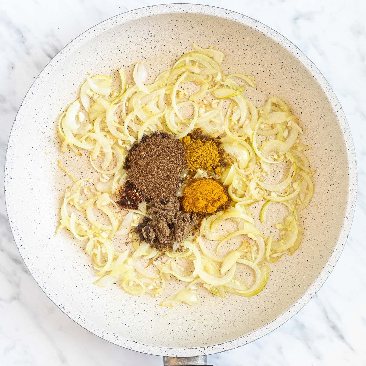 Onion slices with different heaps of spices in a white frying pan. 
