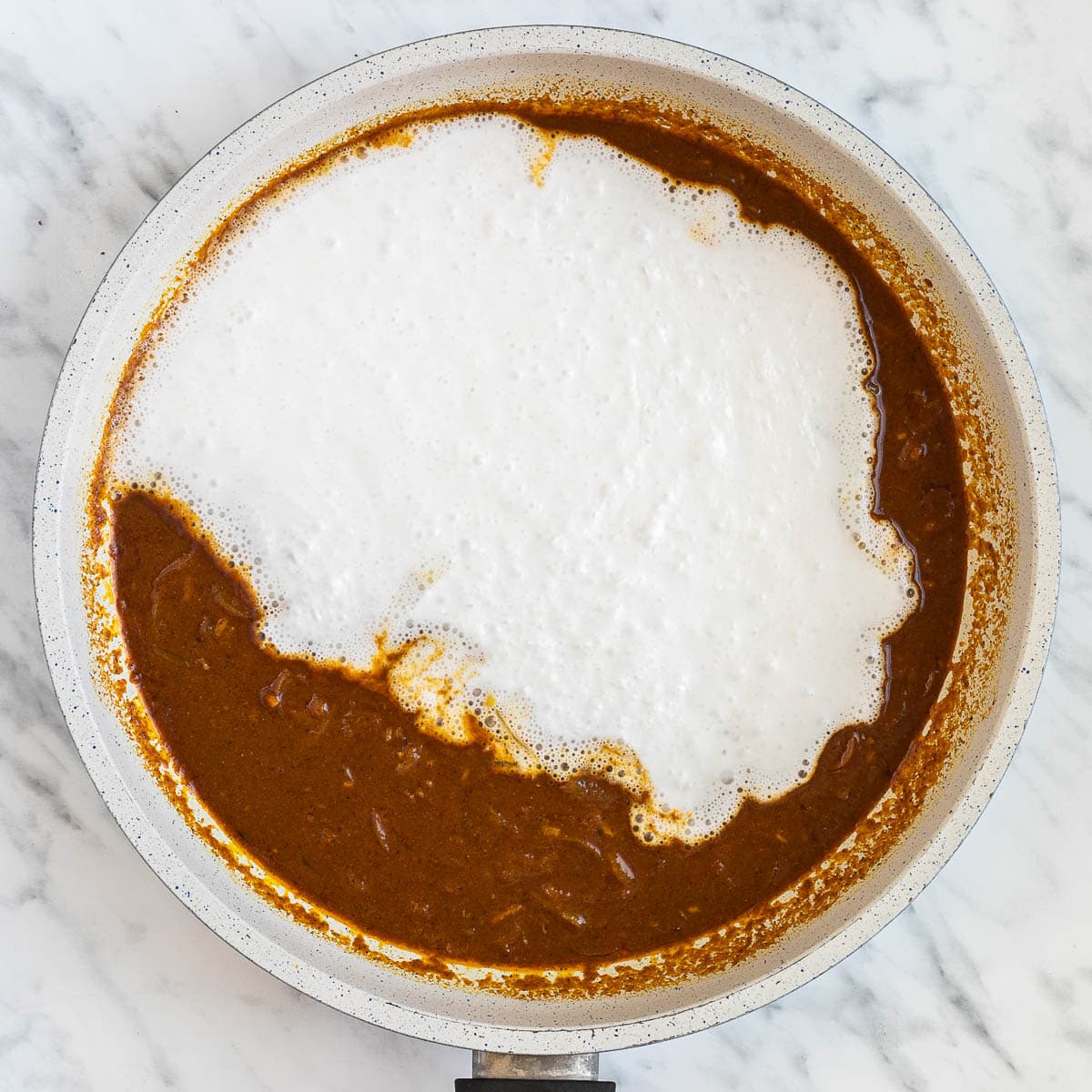 White liquid is added to a dark brown sauce in a white frying pan.