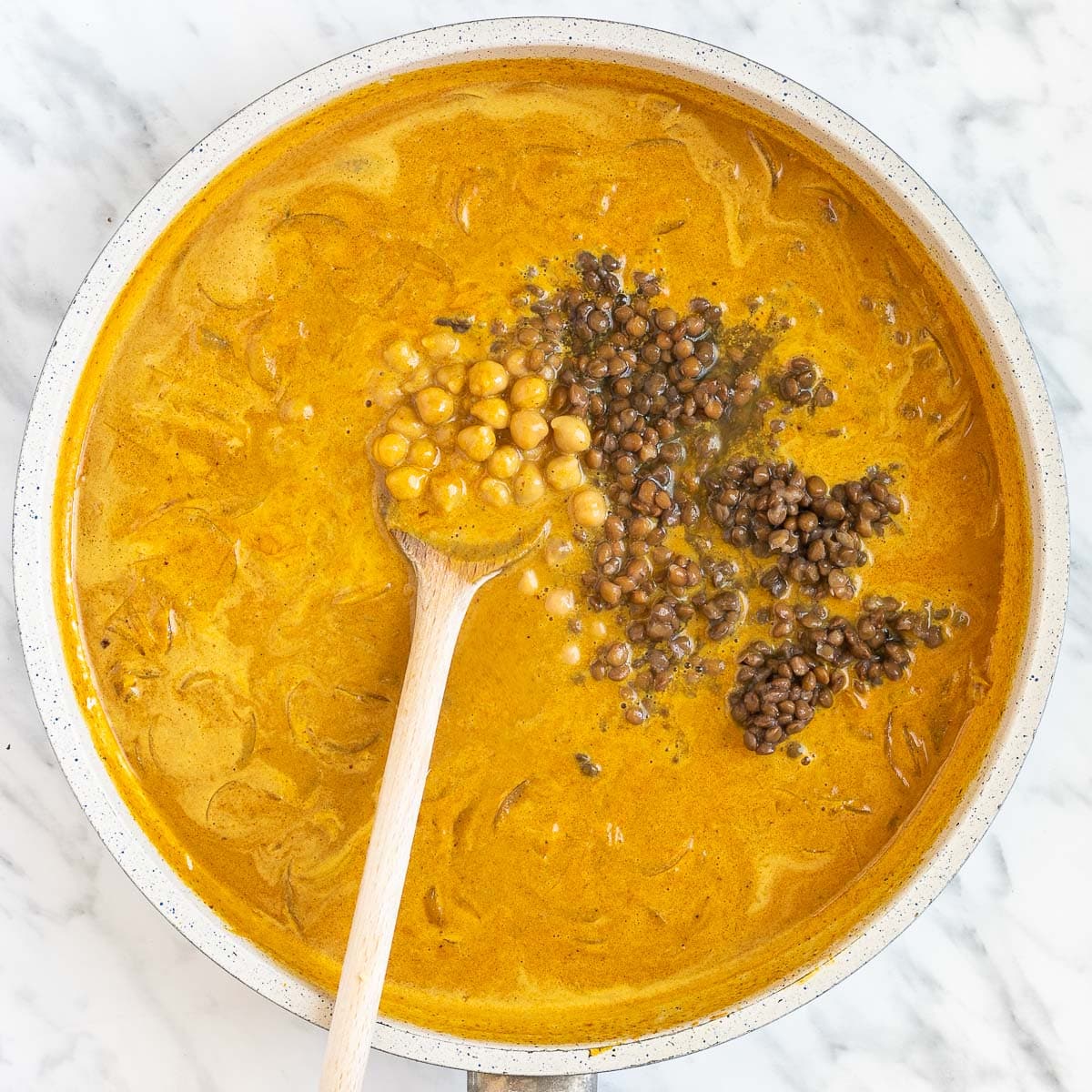 Chickpeas and brown lentils are added with a wooden spoon to an orange light brown sauce in a white frying pan