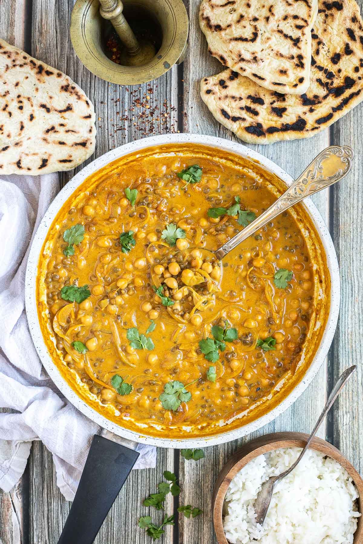 A white frying pan with chickpeas and lentils in a creamy light brown sauce sprinkled freshly chopped cilantro. A spoon is placed inside. Naan bread and a bowl of rice are next to it.