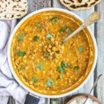 A white frying pan with chickpeas and lentils in a creamy light brown sauce sprinkled freshly chopped cilantro. A spoon is placed inside. Naan bread and a bowl of rice are next to it.