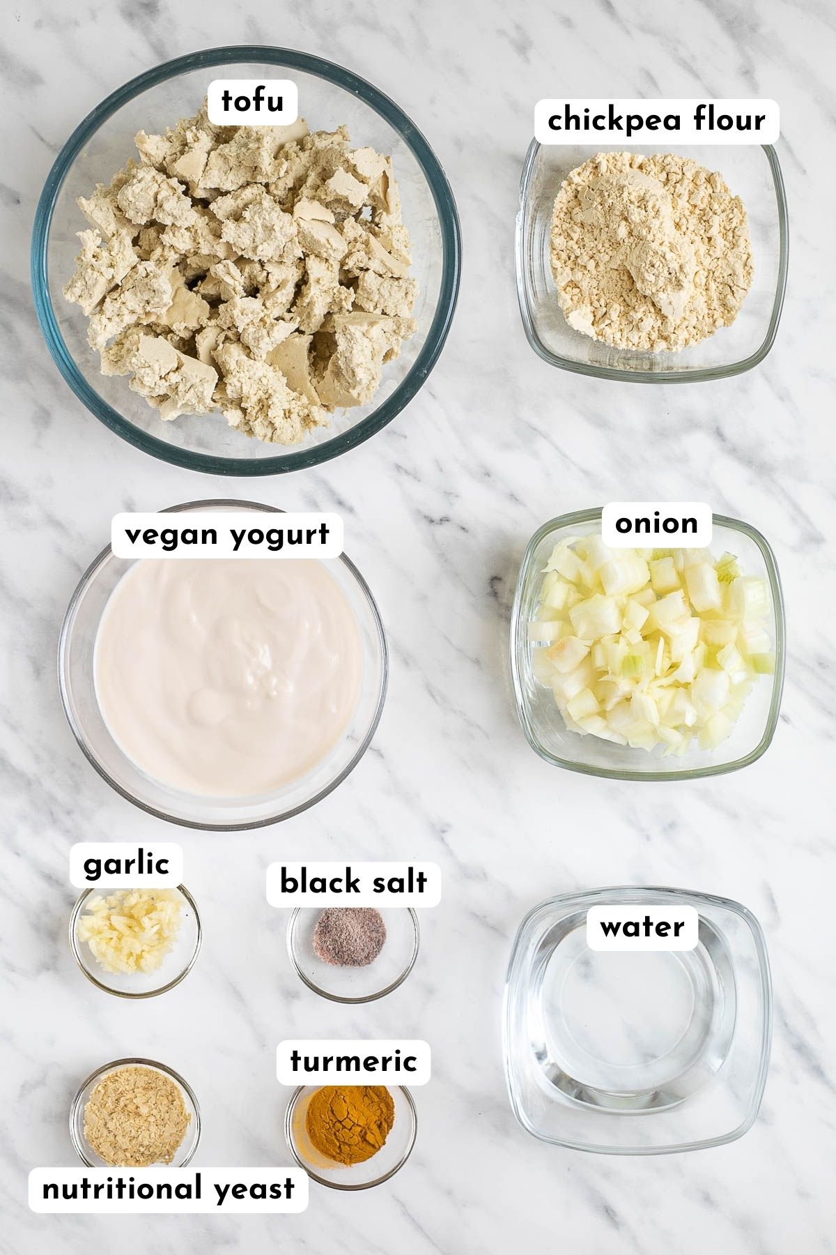 The ingredients of tofu scramble placed in glass bowls like tofu crumbles, chickpea flour, yogurt, onion, garlic, water, yellow flakes and orange spice. 