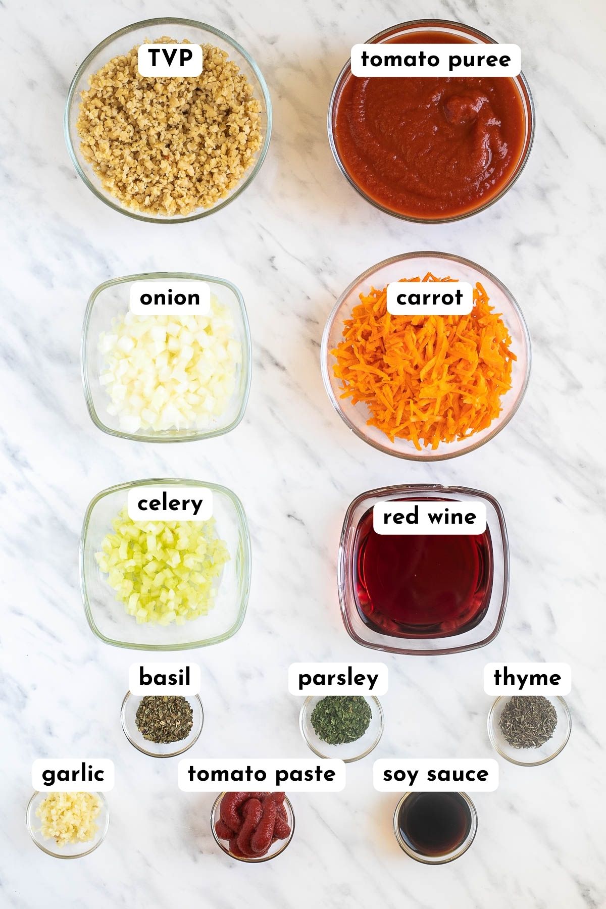 Ingredients of vegan bolognese in small glass containers like plant-based crumbles, tomato sauce, chopped onions, carrot, celery, red wine and several small bowls of spices.