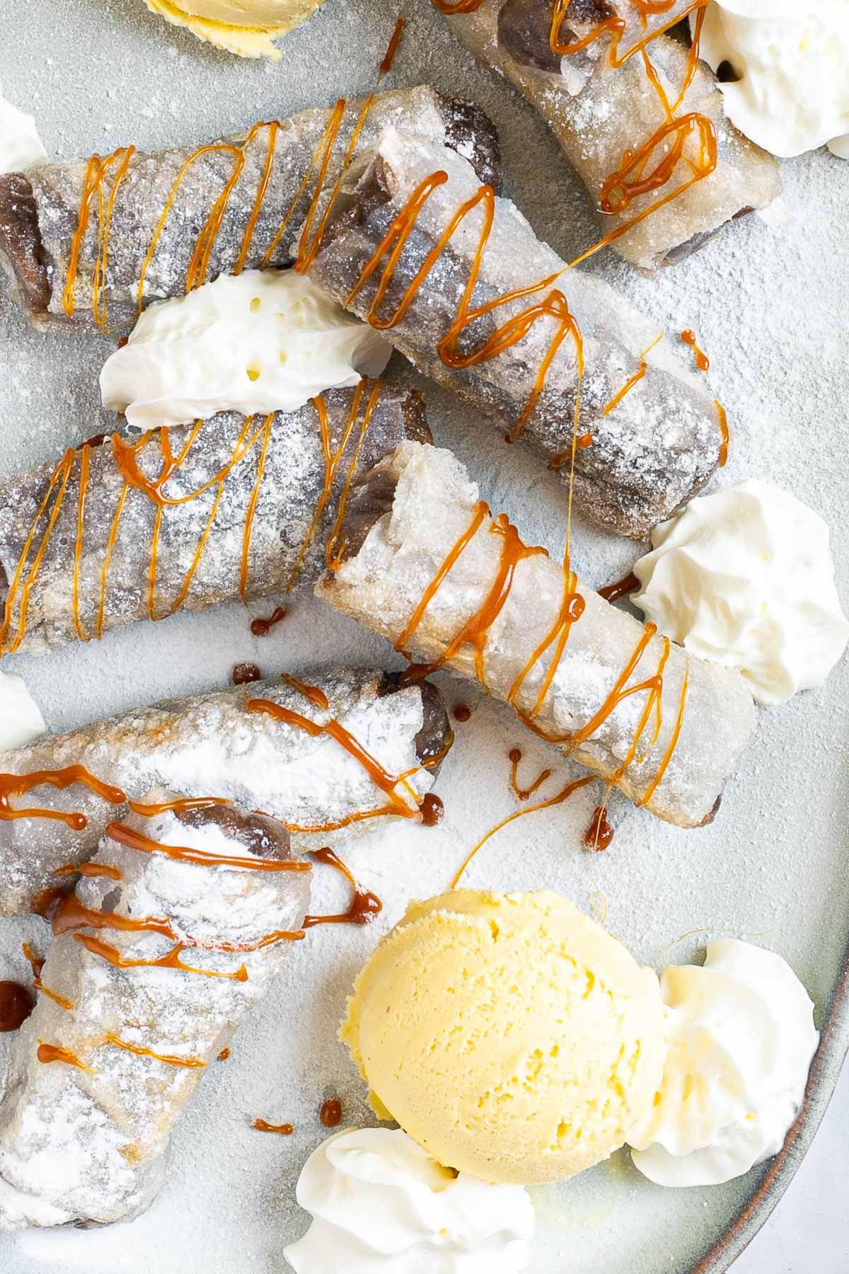 Rice paper rolls with brown filling placed on a light blue plate. Dusted with powdered sugar. Served with scoops of vanilla ice cream, caramel drizzle, and heaps of whipped cream.
