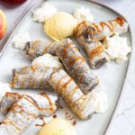 Rice paper rolls with brown filling placed on a light blue plate. Dusted with powdered sugar. Served with scoops of vanilla ice cream, caramel drizzle, and heaps of whipped cream.