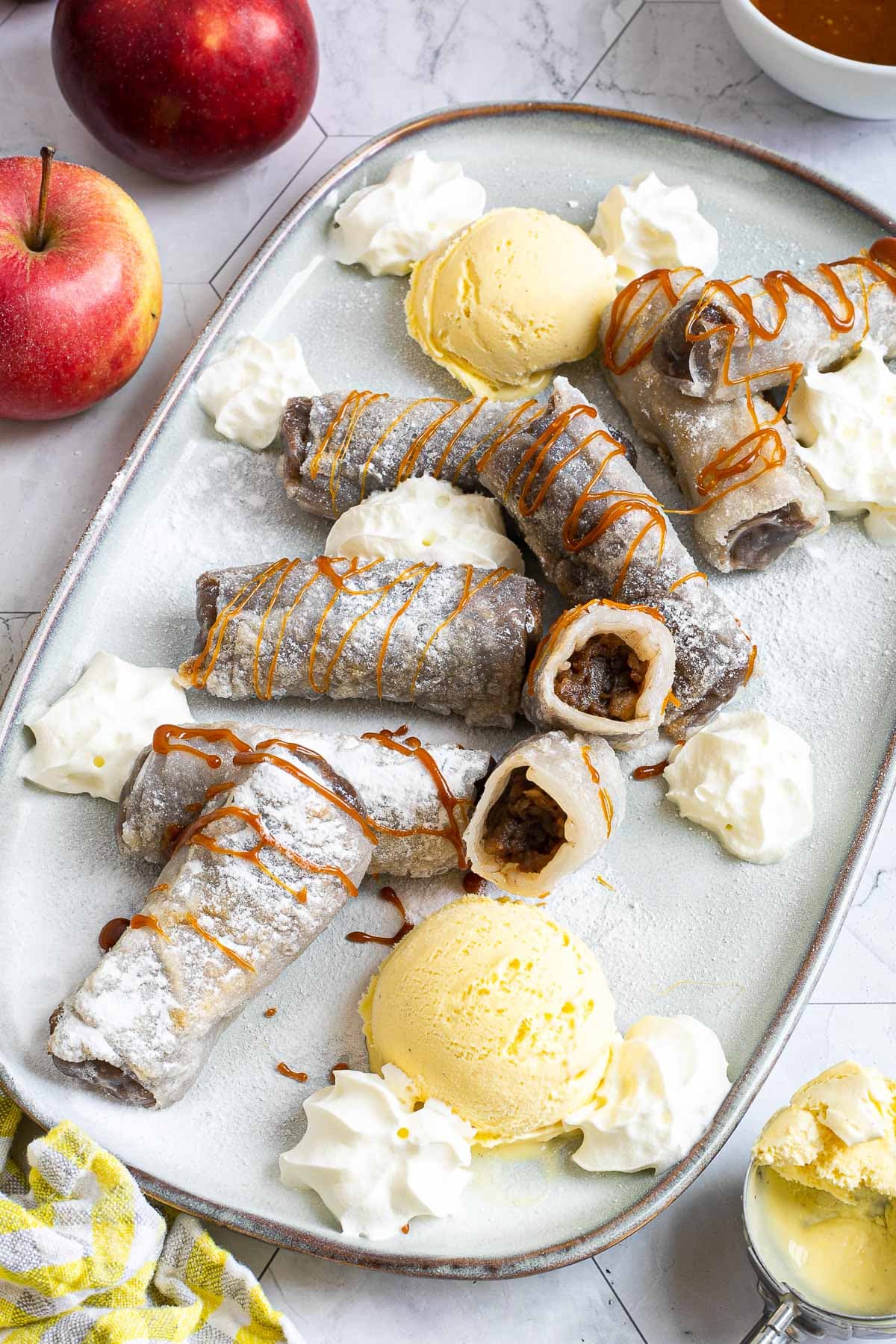 Rice paper rolls with brown filling placed on a light blue plate. Dusted with powdered sugar. Served with scoops of vanilla ice cream, caramel drizzle, and heaps of whipped cream. 2 rolls are cut in half so the filling is visible.