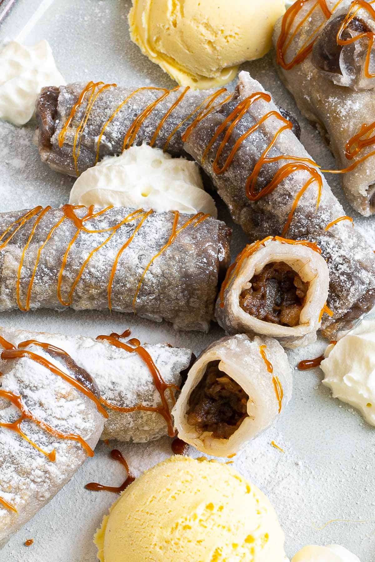 Rice paper rolls with brown filling placed on a light blue plate. Dusted with powdered sugar. Served with scoops of vanilla ice cream, caramel drizzle, and heaps of whipped cream. 2 rolls are cut in half so the filling is visible.
