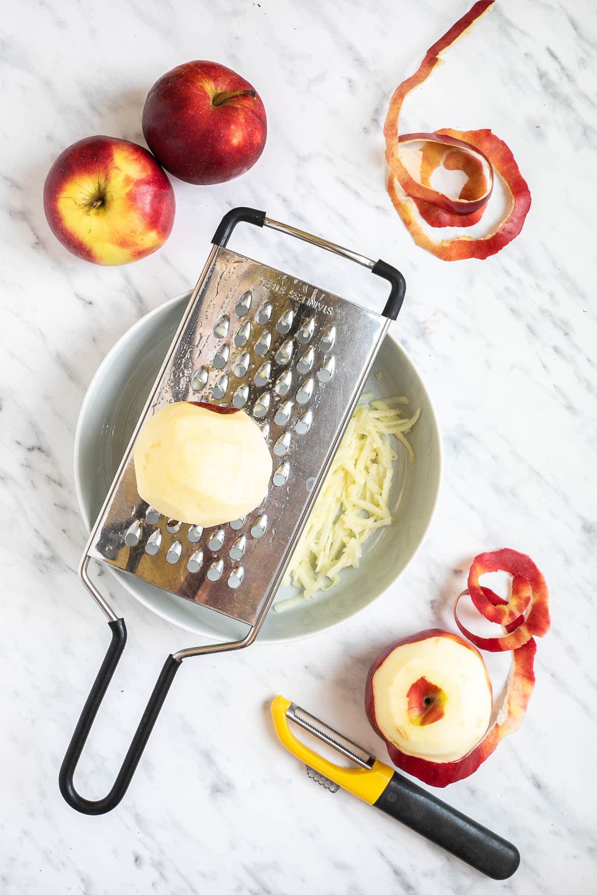 A white bowl with a large hole grater on top. Grated apples are visible in the bowel and a peeled apple is placed on top of the grater. More apples are around it.