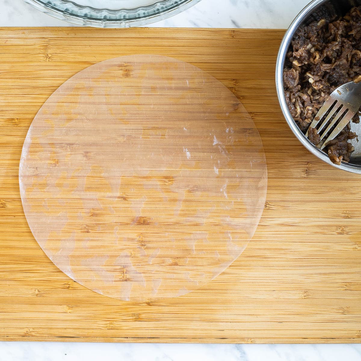 A wooden cutting board with a wet rice paper sheet. Brown grated apples in a small saucepan is next to it.
