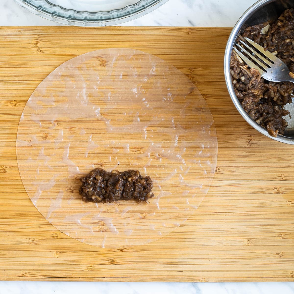 A wooden cutting board with a wet rice paper sheet and a heap of brown grated apple. Brown grated apples in a small saucepan is next to it.