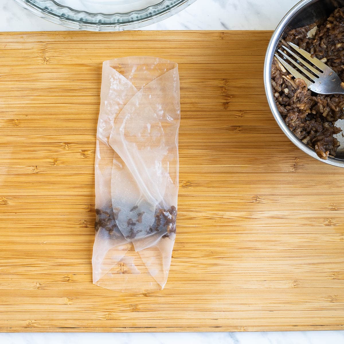A wooden cutting board with a wet rice paper with brown filling folded from the sides. Brown grated apples in a small saucepan is next to it.