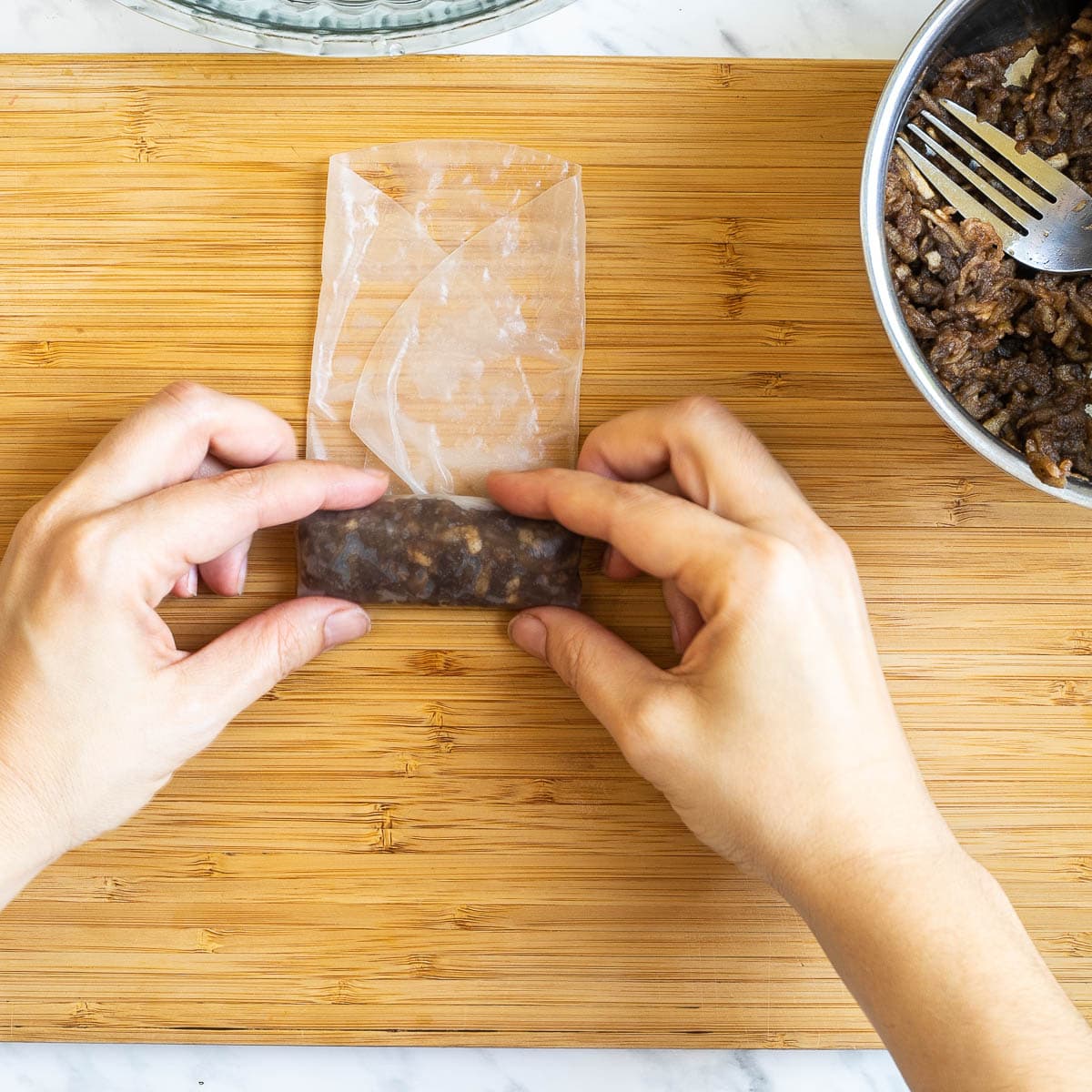A hand is rolling a wet rice paper sheet with a brown filling. Brown grated apples in a small saucepan is next to it.