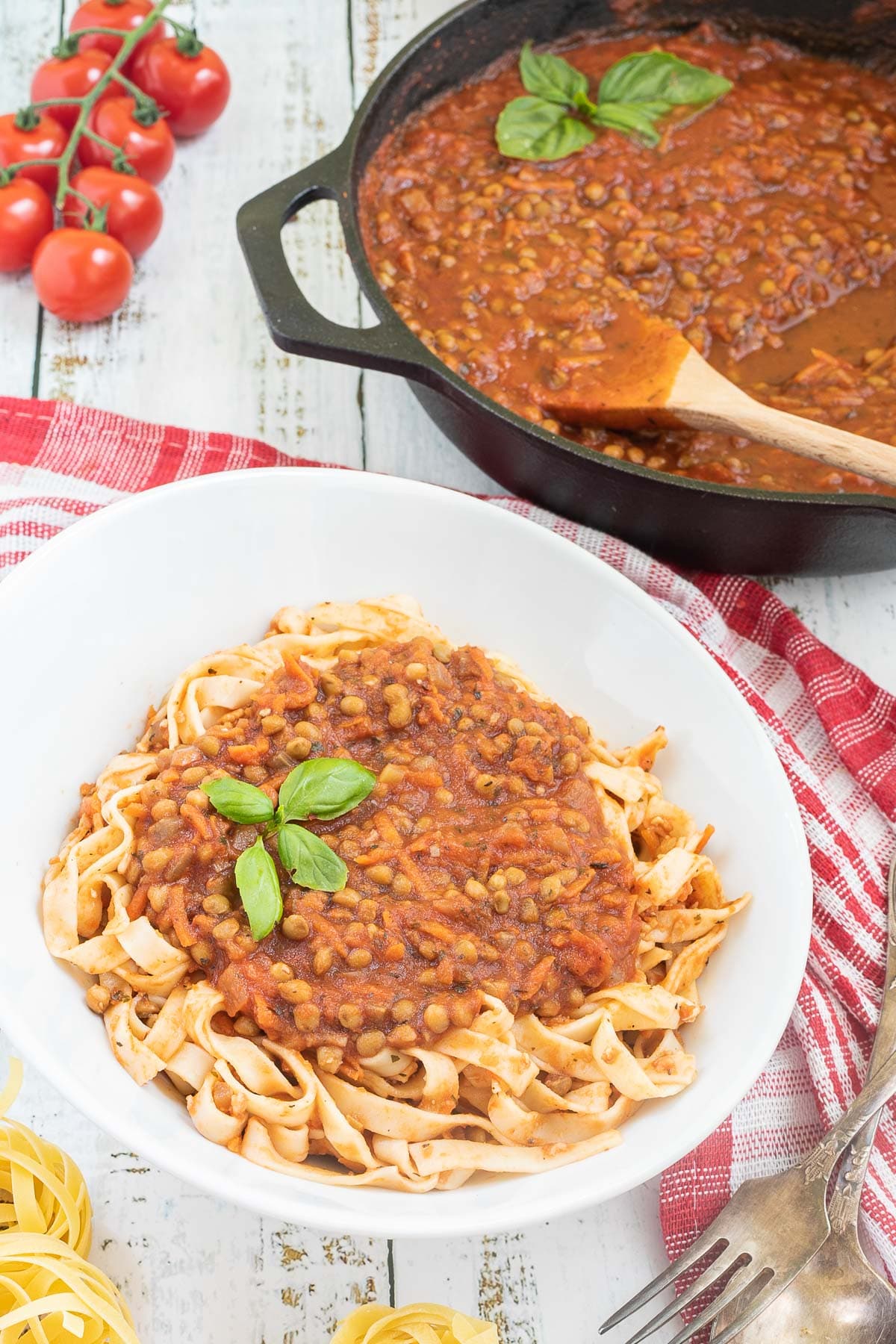 A white bowl of tagliatelle pasta topped with lentils in bolognese sauce. Leftover bolognese in the cast iron skillet is right next to it.
