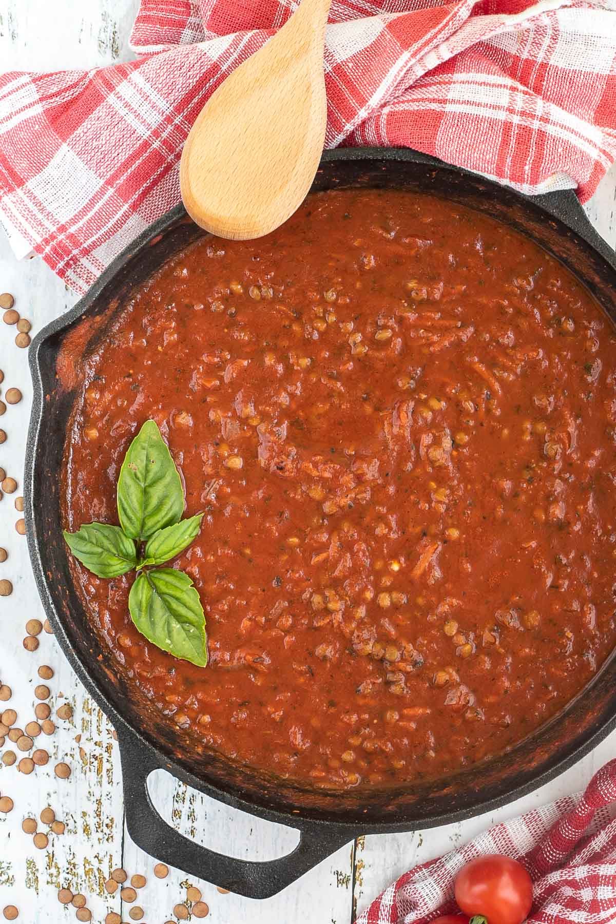 A cast iron skillet with brown lentils in bolognese sauce. Dry lentils and some cherry tomatoes are scattered around the skillet. A wooden spatula is placed on the side.