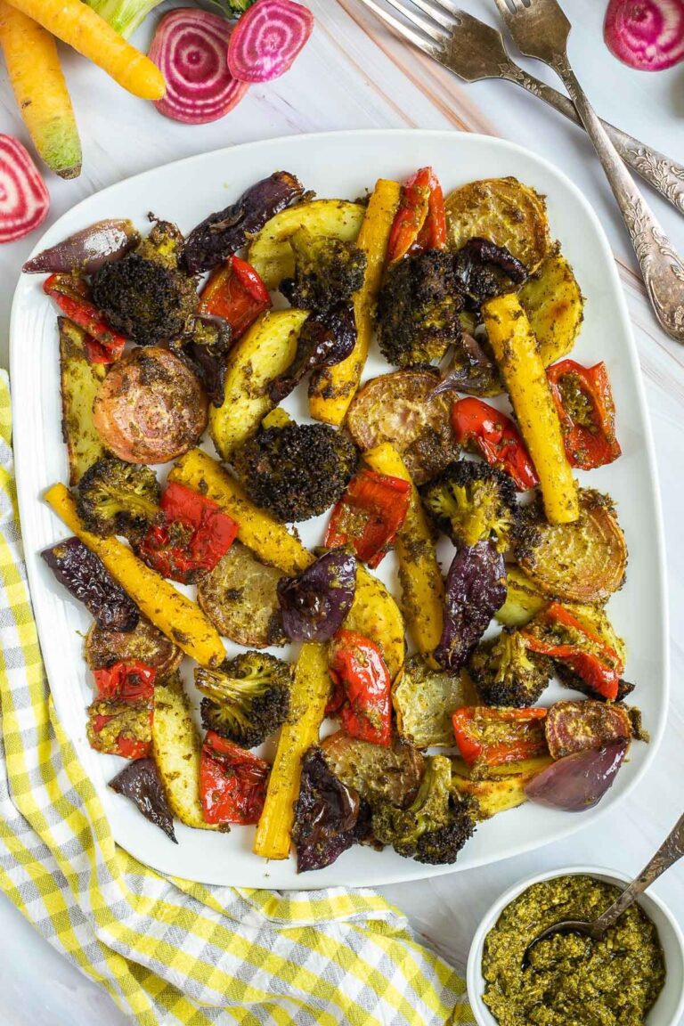 Large white serving plate with vibrant vegetables like broccoli, yellow carrots, red pepper, red onion, pink beet and potato slightly charred around the edges covered in a bit of pesto sauce.