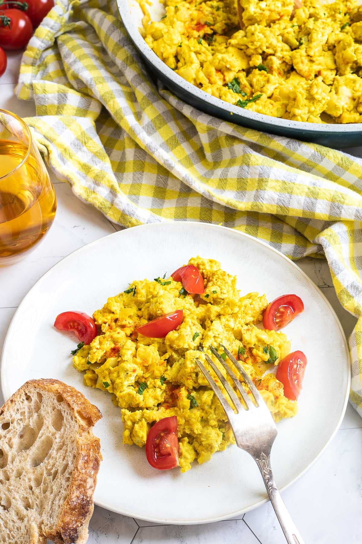 Yellow tofu crumbles and cherry tomato wedges are served on a plate. Remaining crumbles are in a large frying pan. Apple juice and a slice of bread are next to it.