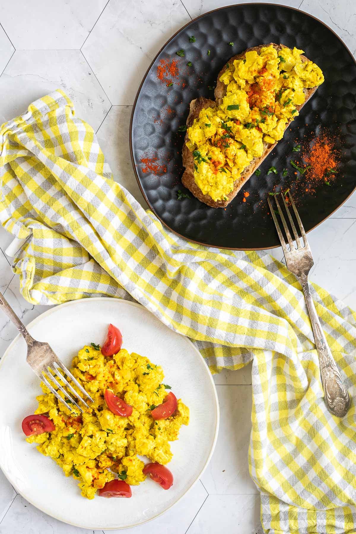 Yellow tofu crumbles and cherry tomato wedges are served on a plate. A slice of bread is topped with the same yellow crumbles is served on a black plate.