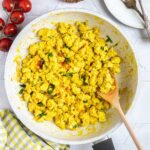 Yellow tofu crumbles in a white frying pan sprinkled with fresh herbs and red pepper flakes. A wooden spatula is placed inside.