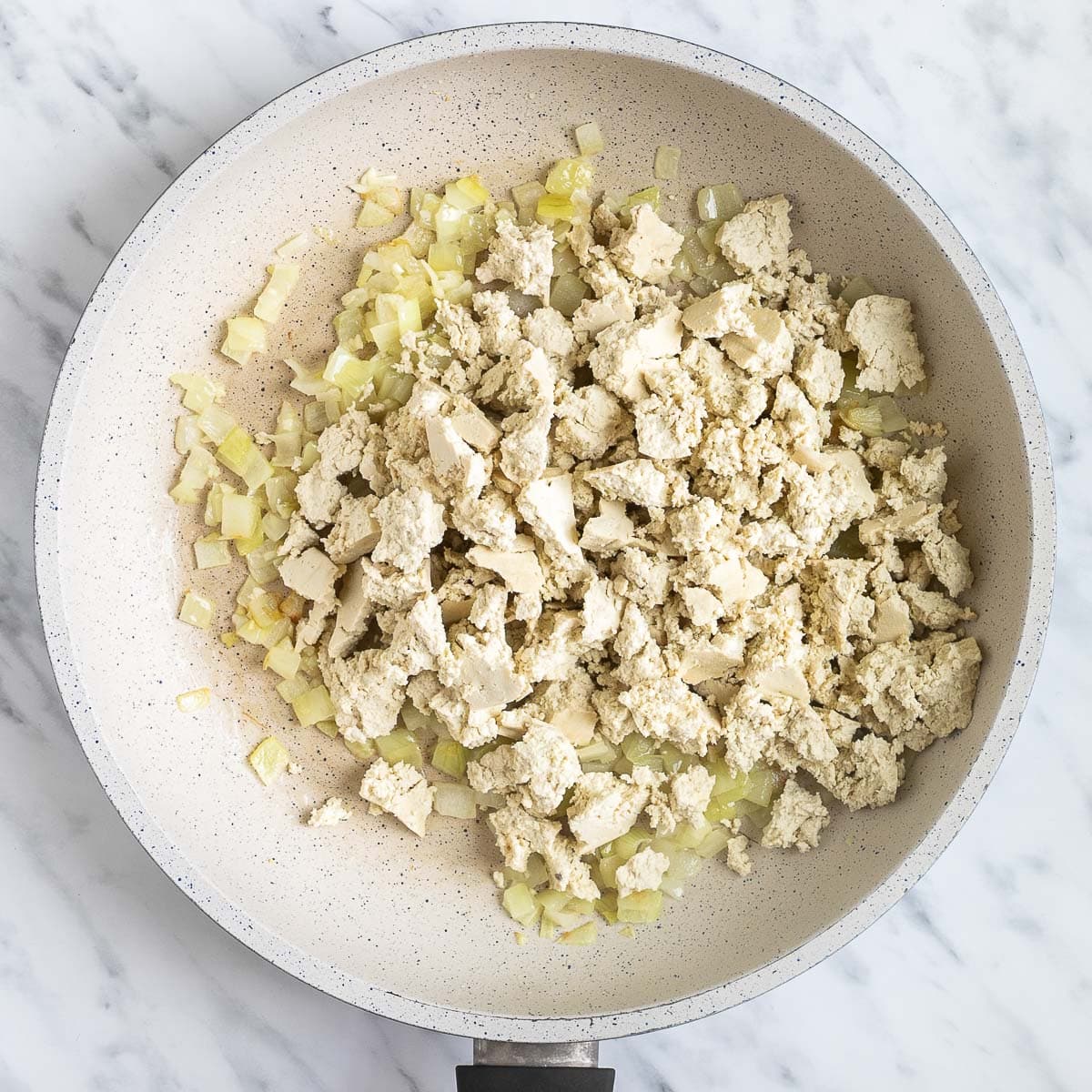 White tofu crumbles and onion, garlic pieces in a white frying pan.
