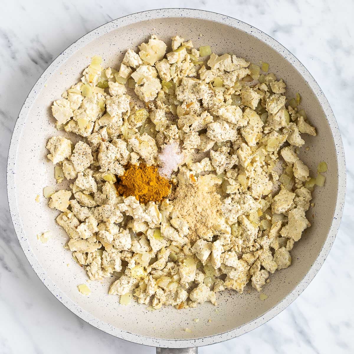 White tofu crumbles, onion, garlic pieces topped with spices in a white frying pan.