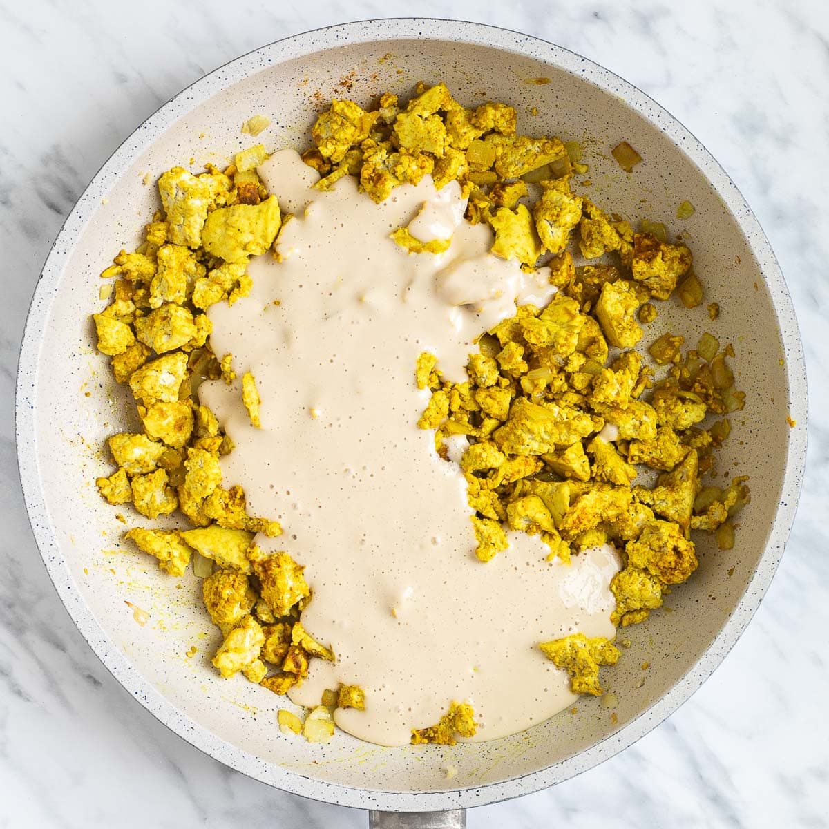 Yellow tofu crumbles, onion, garlic pieces in a white frying pan. A light brown liquid batter is poured in it.