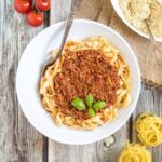 A white bowl of tagliatelle pasta topped with meat-like crumbles in bolognese sauce. Parmesan cheese in a small white bowl right next to it.