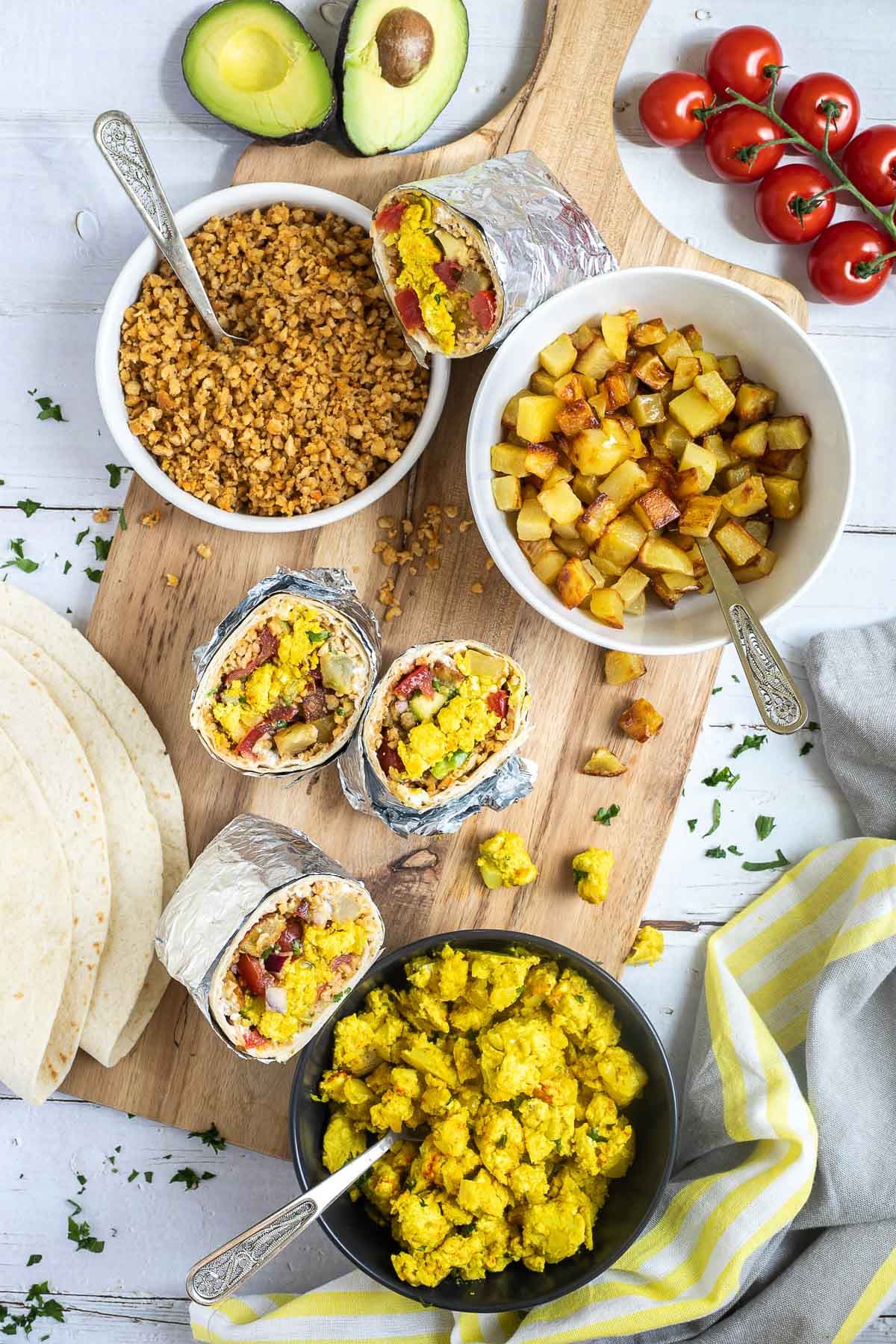 3 half tortillas wrapped in tin foil filled with sausage crumbles, diced potatoes, tofu scrambles, and avocado tomato salsa. The remaining fillings are placed in several bowls on a wooden platter.