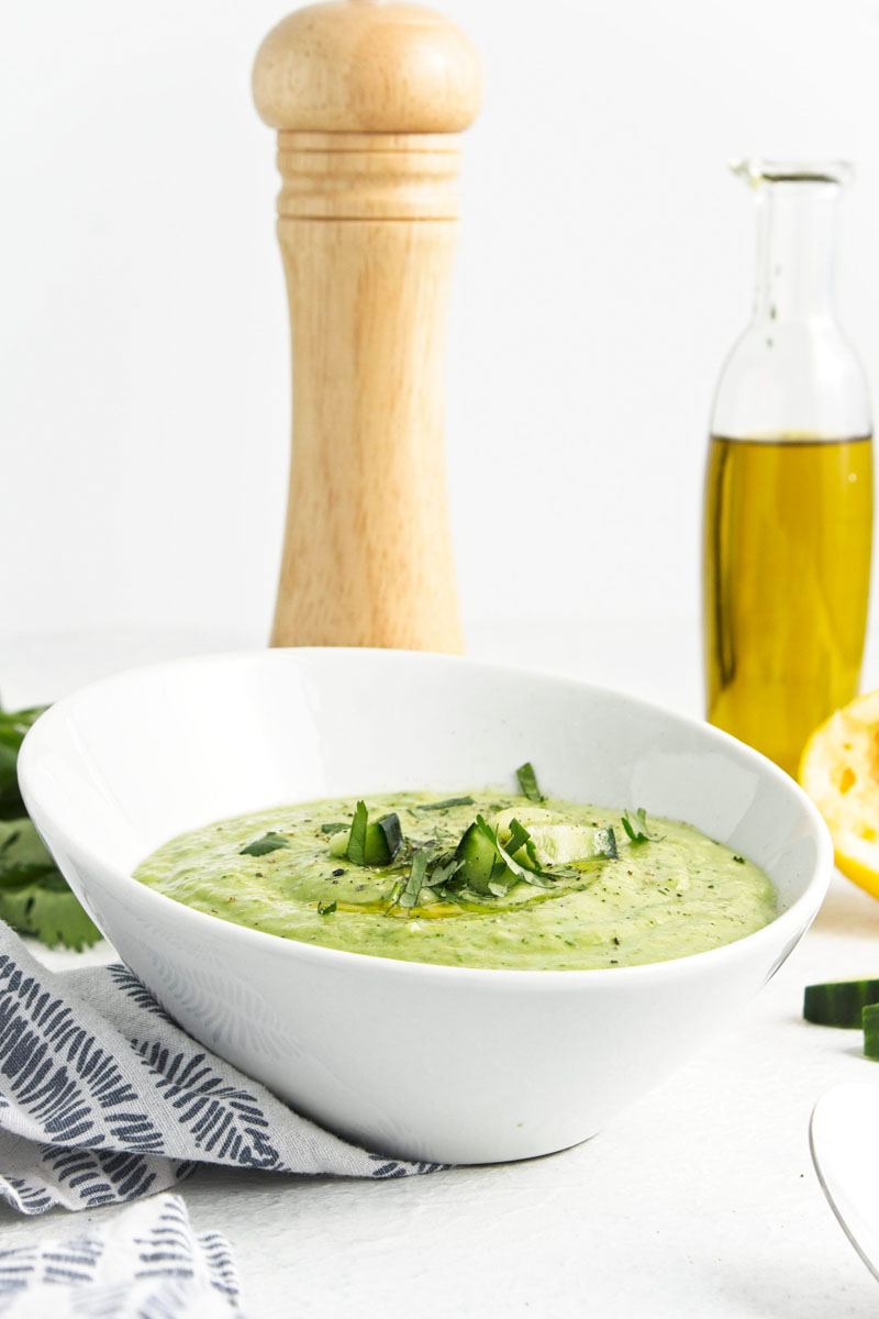 A white bowl with a vibrant green soup topped with freshly chopped herbs and cucumber pieces.