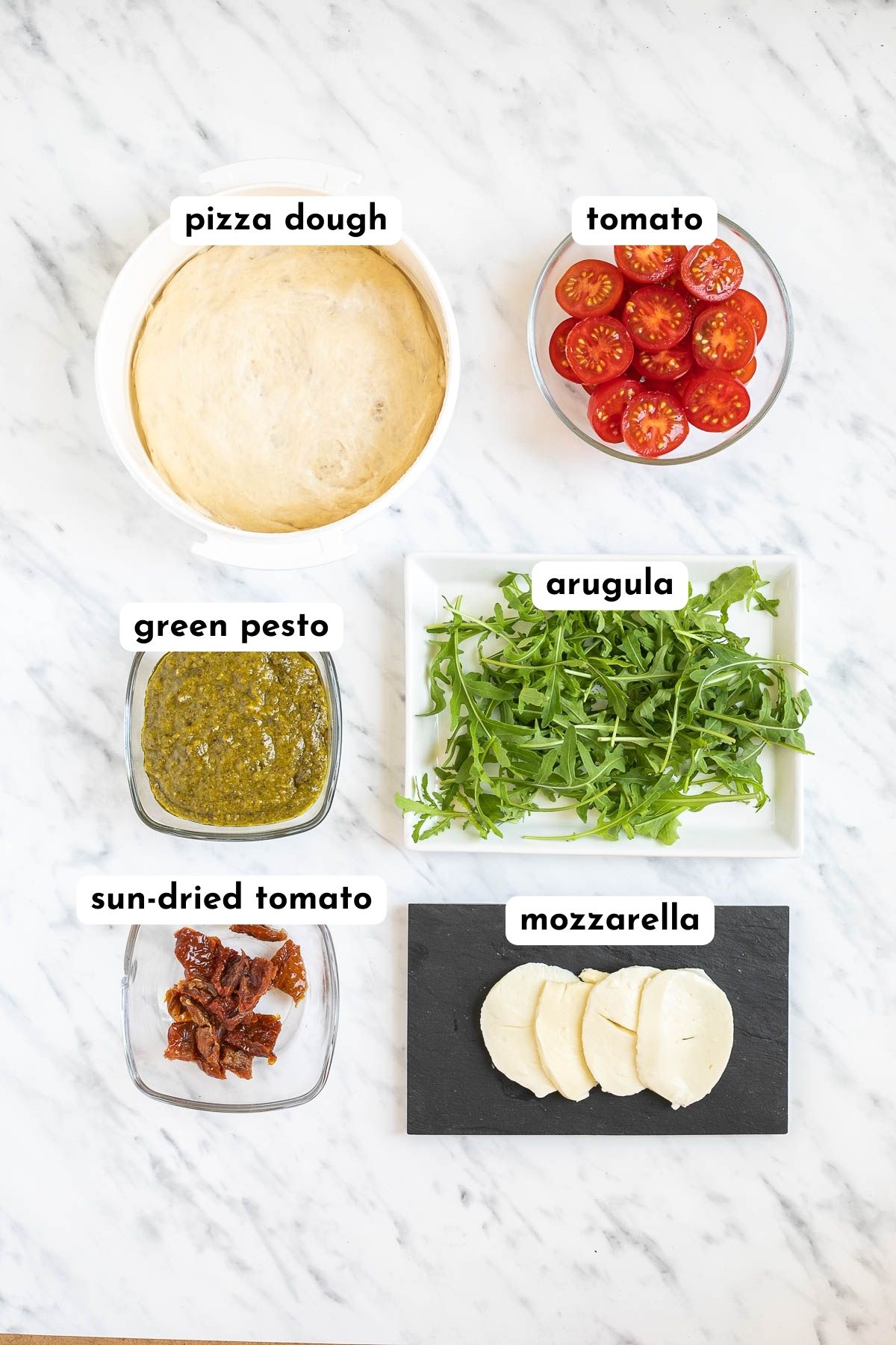 The ingredients of pesto pizza in small containers like pizza dough, cherry tomatoes, sun-dried tomatoes, arugula, pesto, and mozzarella