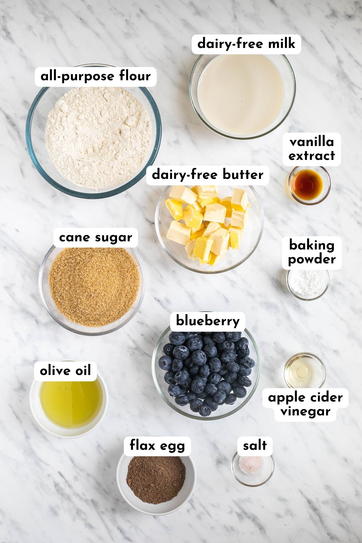 Ingredients of vegan blueberry muffins in small bowls like flour, milk, butter cubes, sugar, oil, vinegar, baking powder, ground flax seeds, salt and fresh blueberries