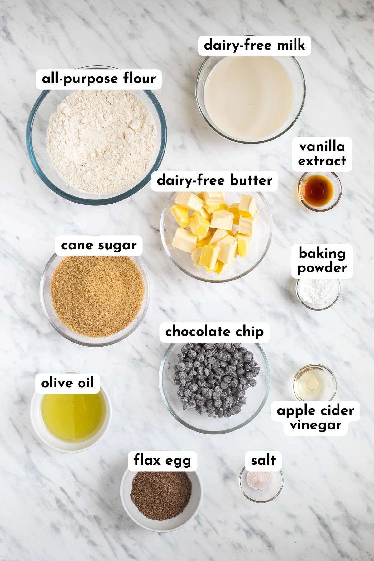 Ingredients of vegan chocolate chip muffins in small bowls like flour, milk, butter cubes, sugar, oil, vinegar, baking powder, ground flax seeds, salt and chocolate chips