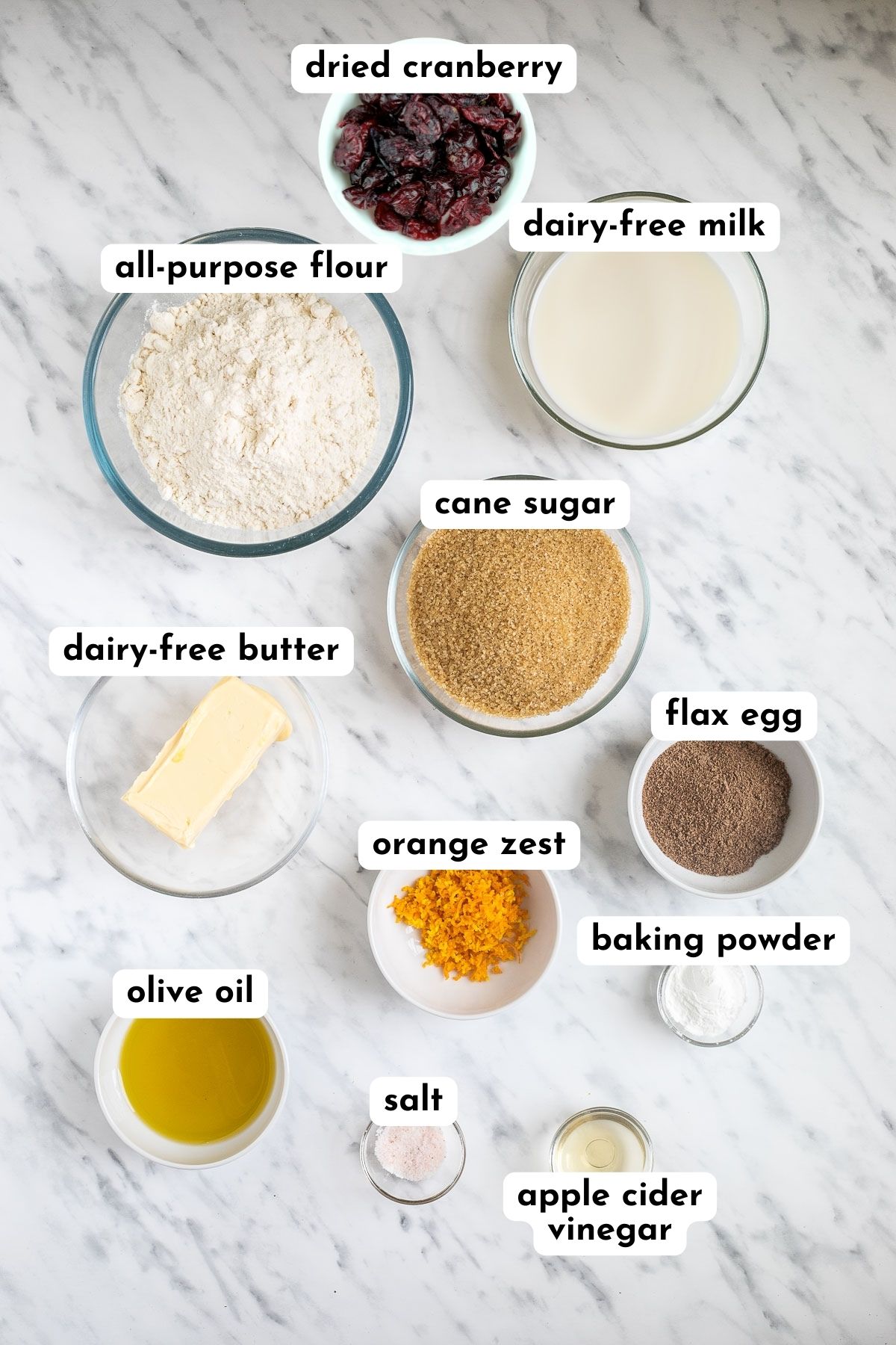 Ingredients of vegan orange cranberry muffins in small bowls like flour, milk, butter cubes, sugar, oil, vinegar, baking powder, ground flax seeds, salt, dried cranberries, and grated orange zest