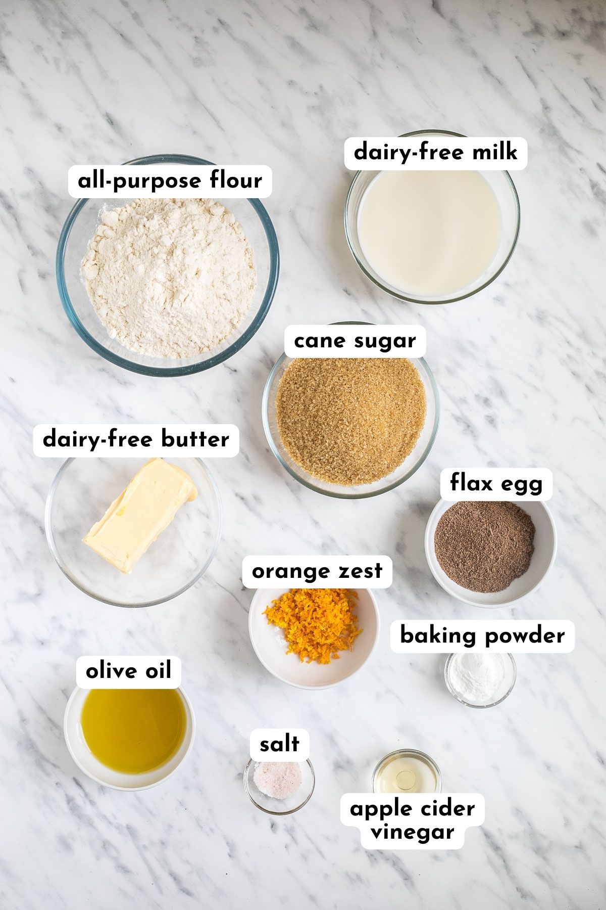 Ingredients of vegan orange cranberry muffins in small bowls like flour, milk, butter cubes, sugar, oil, vinegar, baking powder, ground flax seeds, salt, and grated orange zest