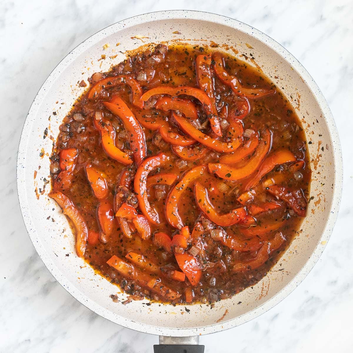 A white frying pan with chopped onions and red pepper slices in a herby tomato sauce.