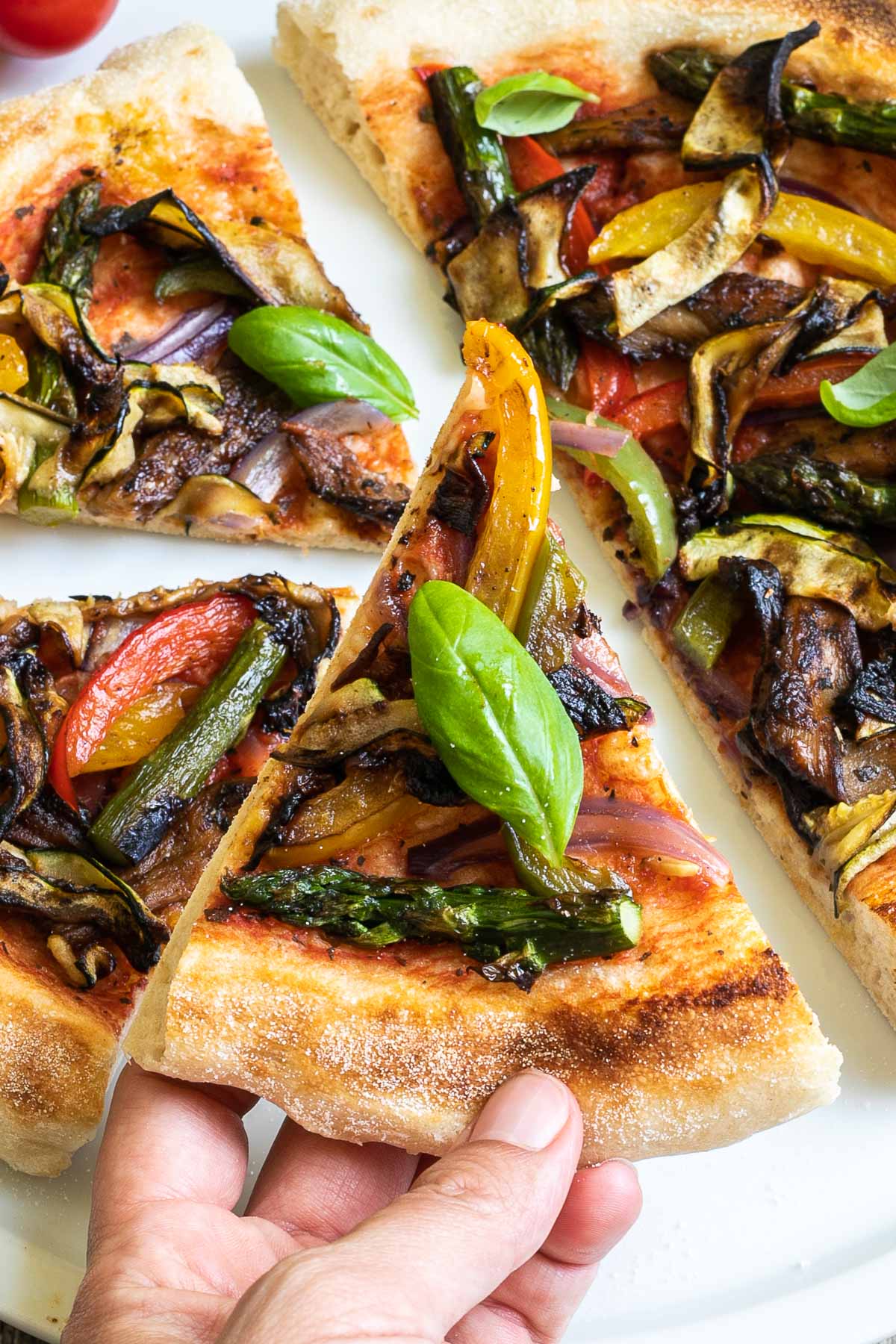 Sliced cheeseless pizza on a white pizza plate topped with tomato sauce, bell pepper strips, zucchini, mushrooms, asparagus, red onion, and basil. A hand is holding one slice.