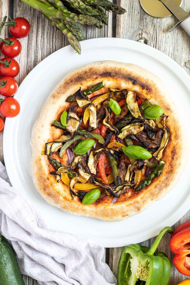 Cheeseless pizza on a white pizza plate topped with tomato sauce, bell pepper strips, zucchini, mushrooms, asparagus, red onion, and basil. More fresh ingredients are next to it.