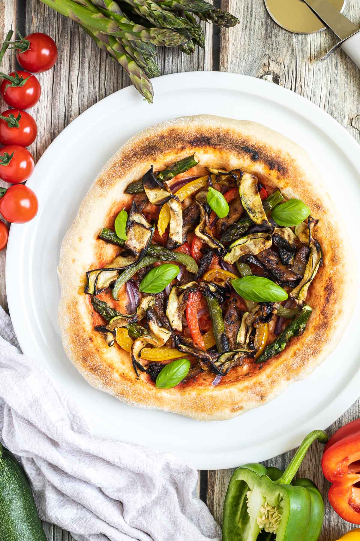 Cheeseless pizza on a white pizza plate topped with tomato sauce, bell pepper strips, zucchini, mushrooms, asparagus, red onion, and basil. More fresh ingredients are next to it. 